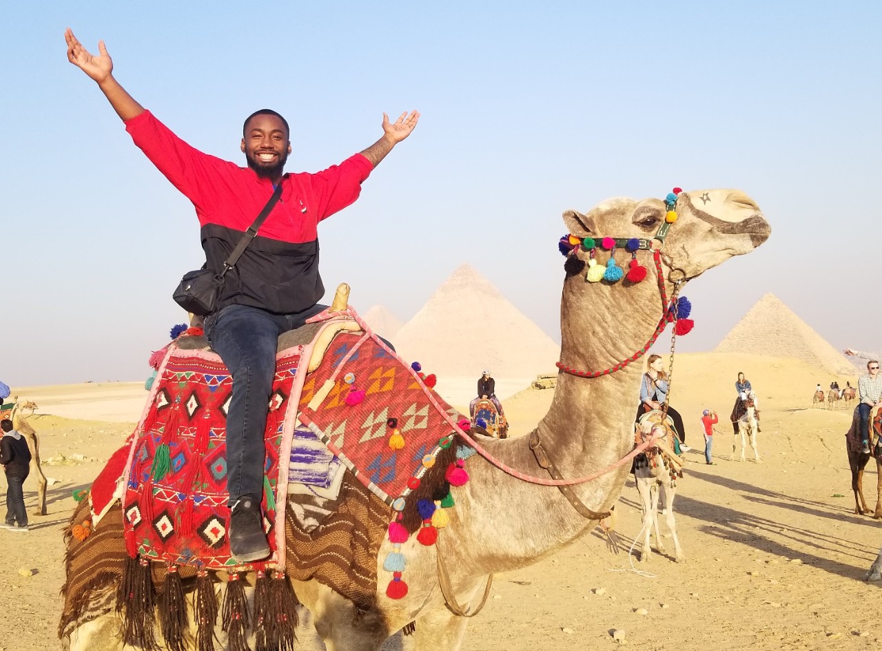 Quoran Knights holds his arms up while riding on a camel in front of the Great Pyramids of Giza in Egypt.